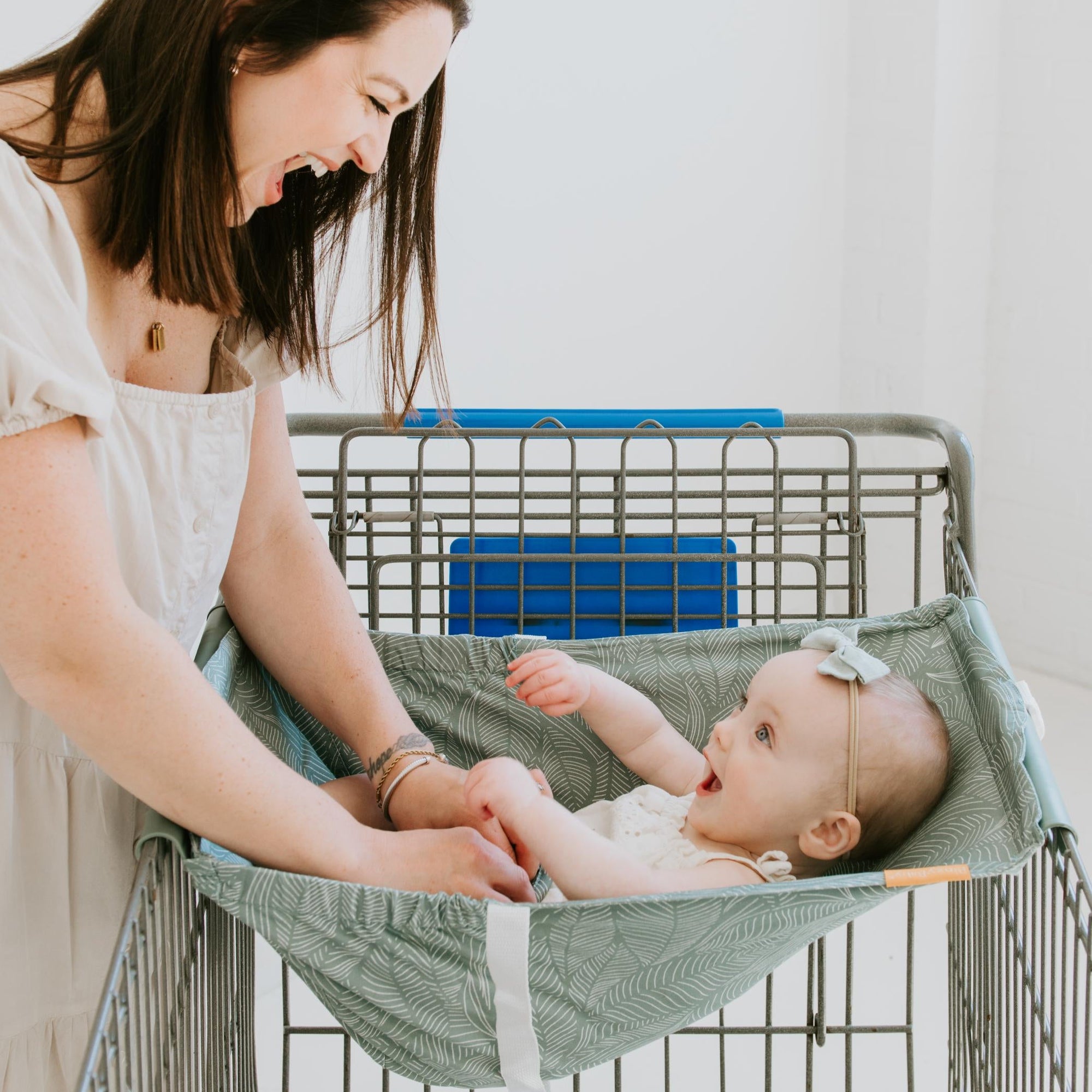 Woman holding a baby in a green binxy baby hammock attached to a shopping cart.