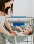 Woman holding a baby in a green binxy baby hammock attached to a shopping cart.